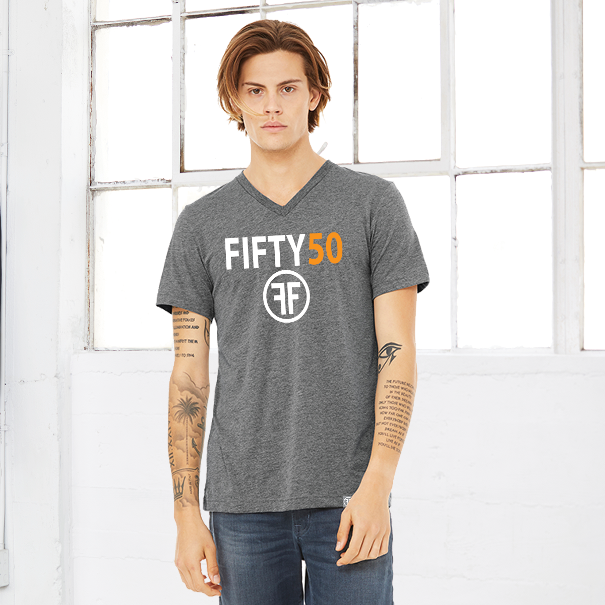 A man wearing a heather grey v-neck t-shirt with a logo. The text Fifty is in white bold font and the number fifty is in orange bold font. Under the text are two white Fs, back to back, surrounded by a circle. 