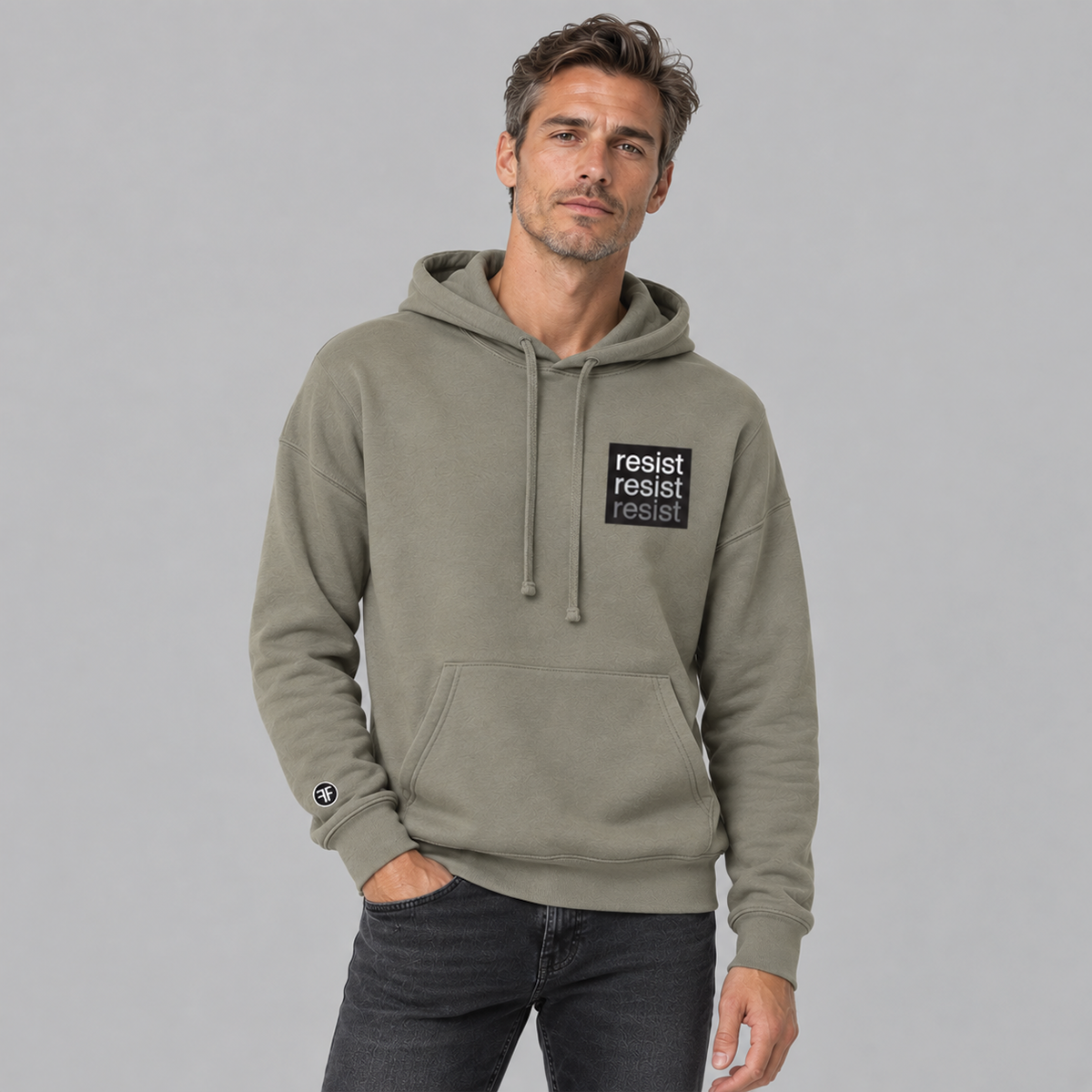 A man wearing a stone taupe pullover hoodie with a black rectangular chest patch that reads 'resist resist resist' in white. light grey and dark grey text, paired with dark gray jeans, against a solid gray studio background.