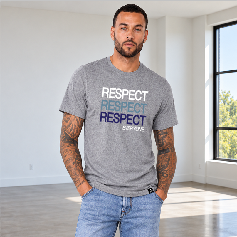 A man wearing a heather gray graphic t-shirt with the word 'RESPECT' printed in white, light blue, and dark blue, paired with light blue jeans, standing in a bright, empty room.