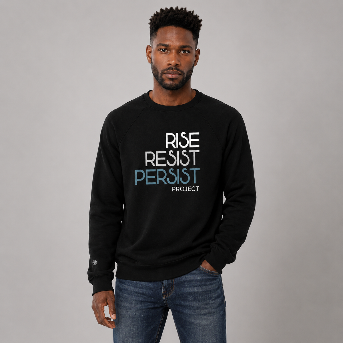 A man wearing a black crewneck sweatshirt with the text 'RISE RESIST PERSIST PROJECT' printed on the chest in white and light blue, paired with blue jeans, against a solid gray studio background.