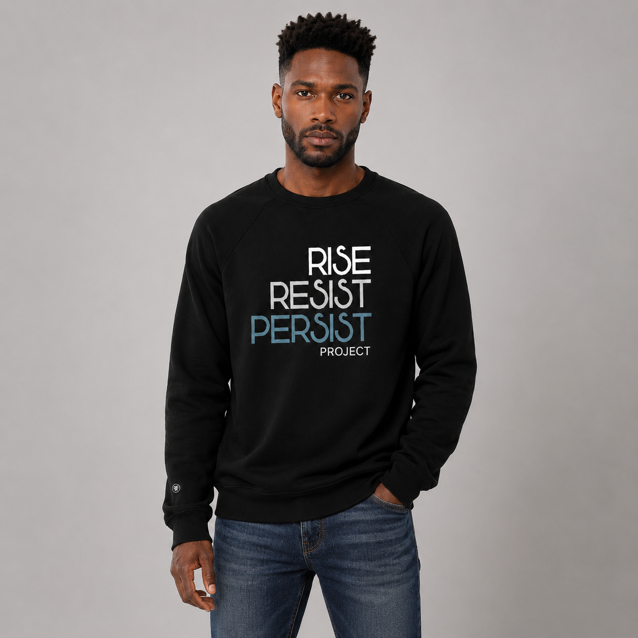 A man wearing a black crewneck sweatshirt with the text 'RISE RESIST PERSIST PROJECT' printed on the chest in white and light blue, paired with blue jeans, against a solid gray studio background.