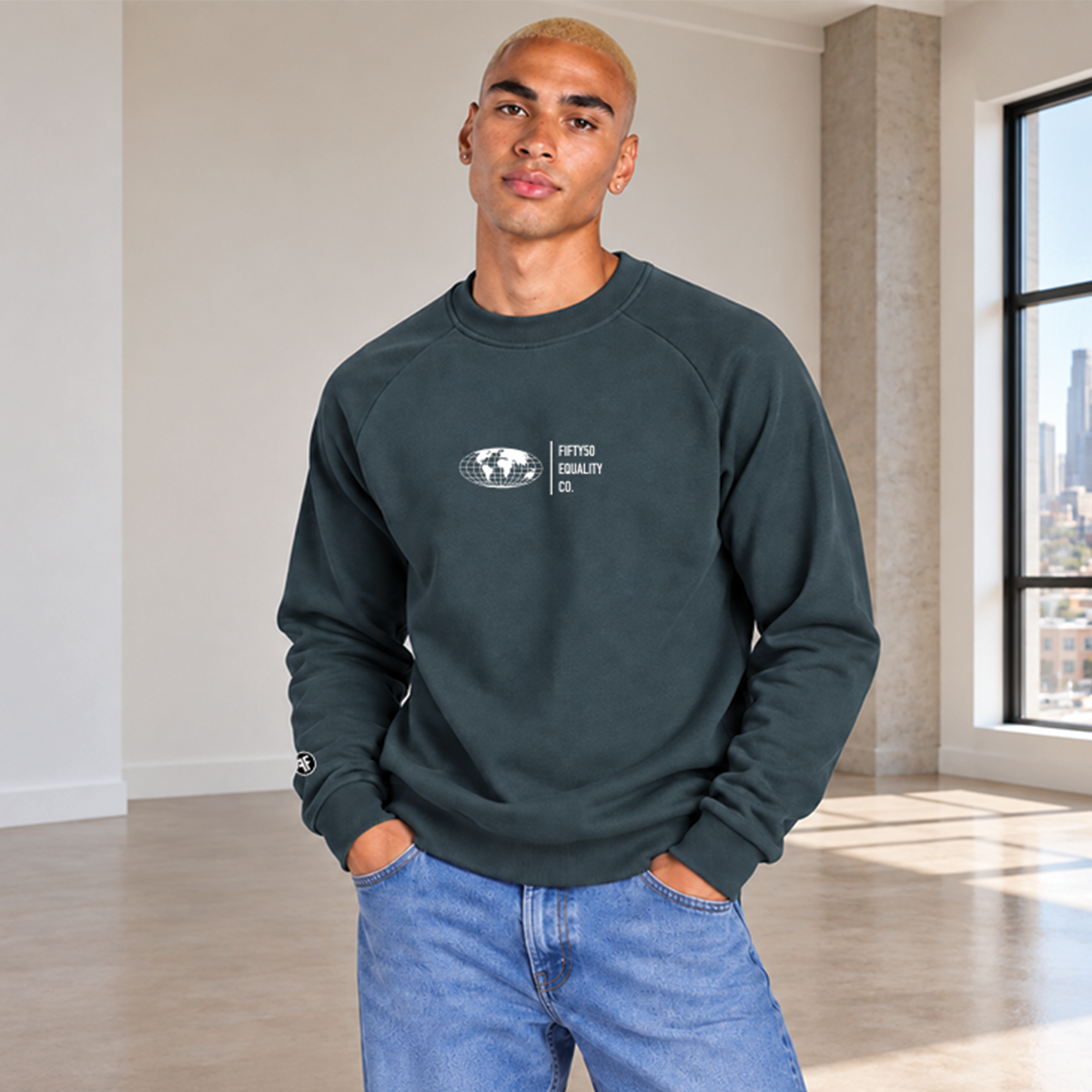 A man wearing a slate teal crewneck sweatshirt with a small white graphic on the front center chest . The graphic features a globe and the words 'FIFTY50 EQUALITY CO.'