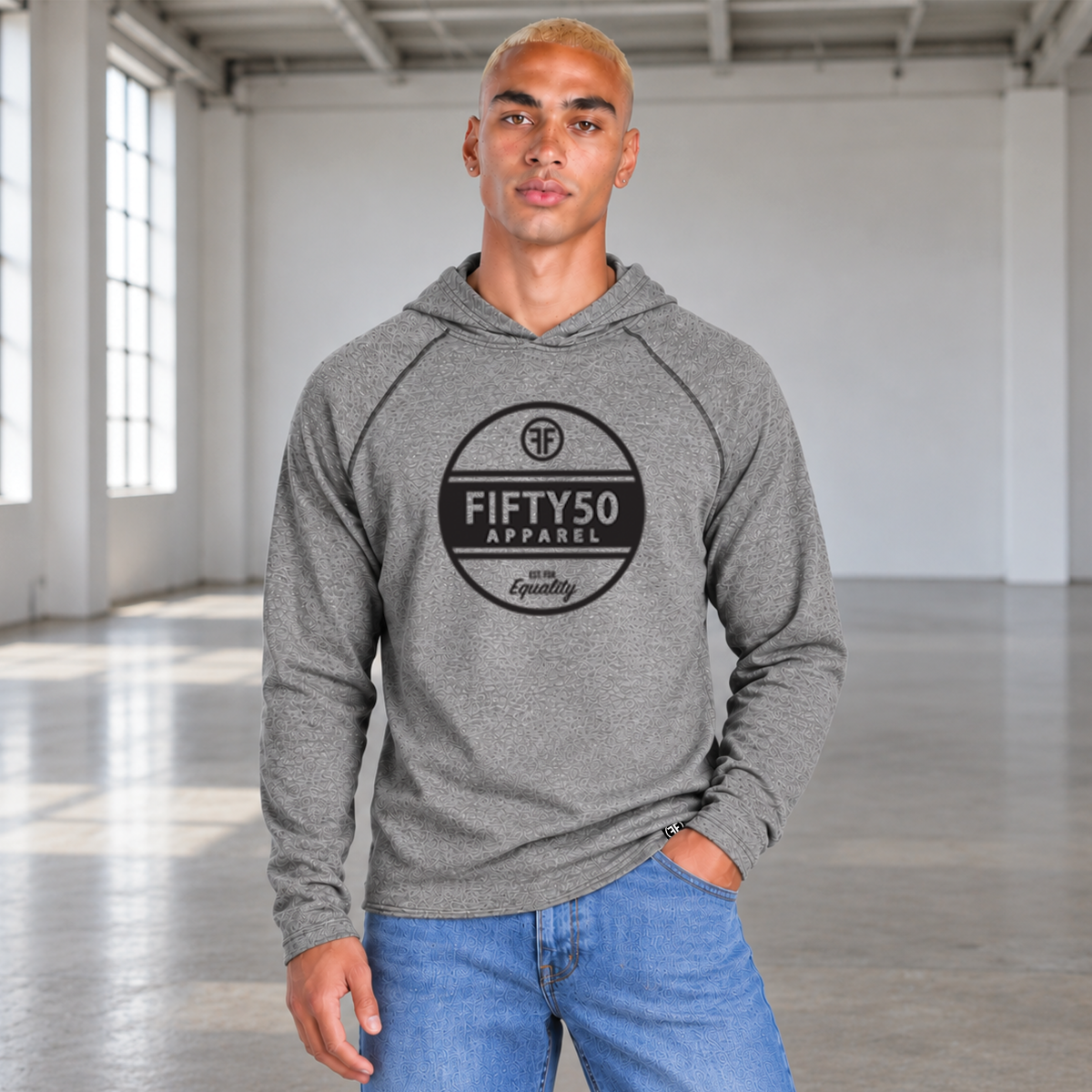 A man wearing a heather gray pullover jersey hoodie with a black circular graphic center chest , that reads 'FIFTY50 APPAREL EST FOR EQUALITY' paired with blue jeans, standing in a bright, modern industrial loft.