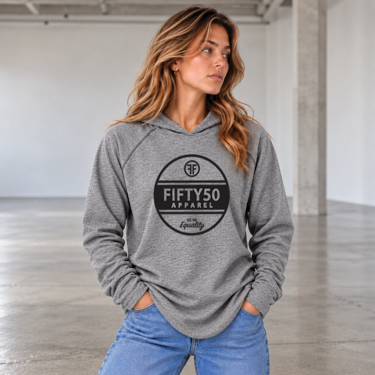 A man wearing a heather gray pullover jersey hoodie with a black circular graphic center chest , that reads 'FIFTY50 APPAREL EST FOR EQUALITY' paired with blue jeans, standing in a bright, modern industrial loft.