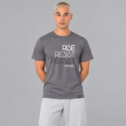 A man wearing a heather slate purple graphic t-shirt with the words 'RISE RESIST PERSIST' and 'PROJECT' printed on the front in white, light grey, and charcoal grey, paired with light blue jeans, set against a solid gray background.
