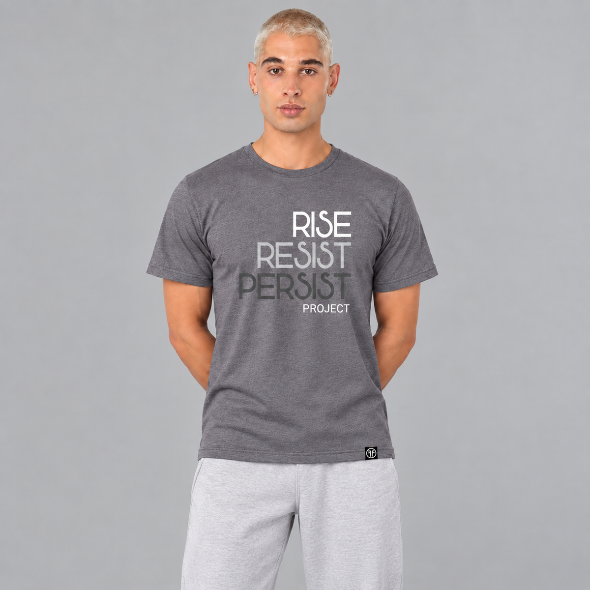 A man wearing a heather slate purple graphic t-shirt with the words 'RISE RESIST PERSIST' and 'PROJECT' printed on the front in white, light grey, and charcoal grey, paired with light blue jeans, set against a solid gray background.