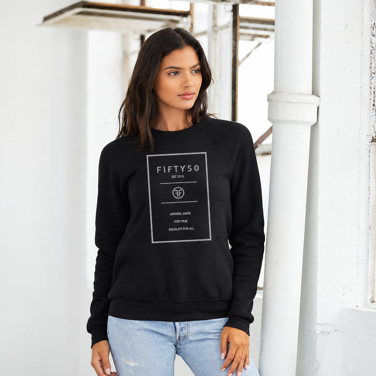 A woman wearing a black crew sweatshirt. The logo is a grey rectangle with the grey text 'Fifty 50', and 'Apparel Made For True Equality for All' inside of it.