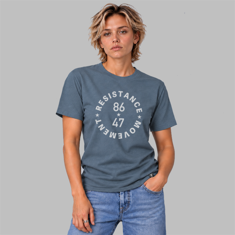 A woman modeling a slate green crewneck t-shirt with a light grey circular graphic that reads 'RESISTANCE MOVEMENT 86 47' with three stars in the logo. She is wearing blue jeans and standing against a solid gray studio background.
