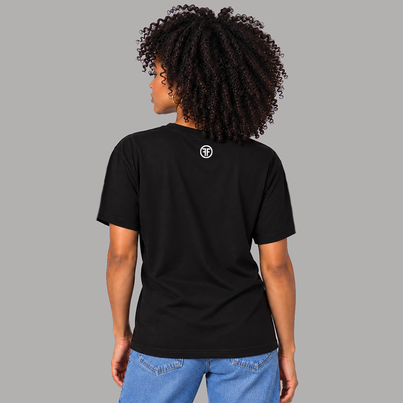 A woman with curly dark hair is shown from the back wearing a black short-sleeve t-shirt to show the unisex fit and that there is a small white circular logo on the upper back, paired with blue jeans, against a solid gray background.
