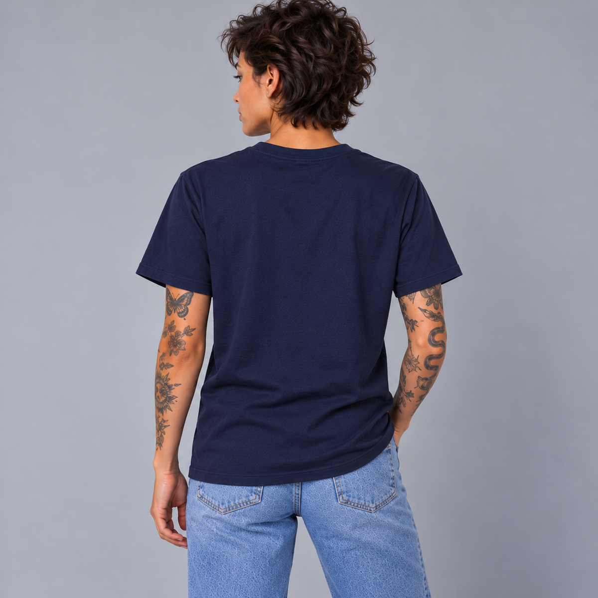 A woman with short dark hair and tattoos, shown from the back wearing a navy blue crewneck t-shirt showing a unisex fit and that there are no graphics on the tee, paired with light blue jeans, against a solid gray studio background.