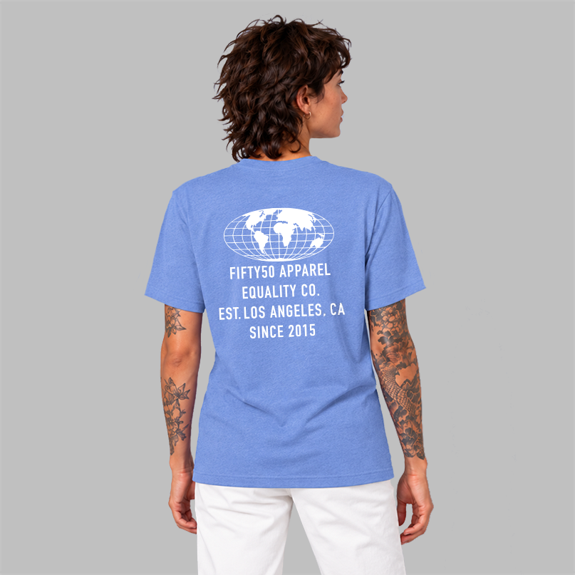 The back of a woman wearing a blue short-sleeve t-shirt with a large white graphic featuring a globe and the text 'FIFTYT50 EQUALITY CO. Paired with white pants, set against a solid gray background.