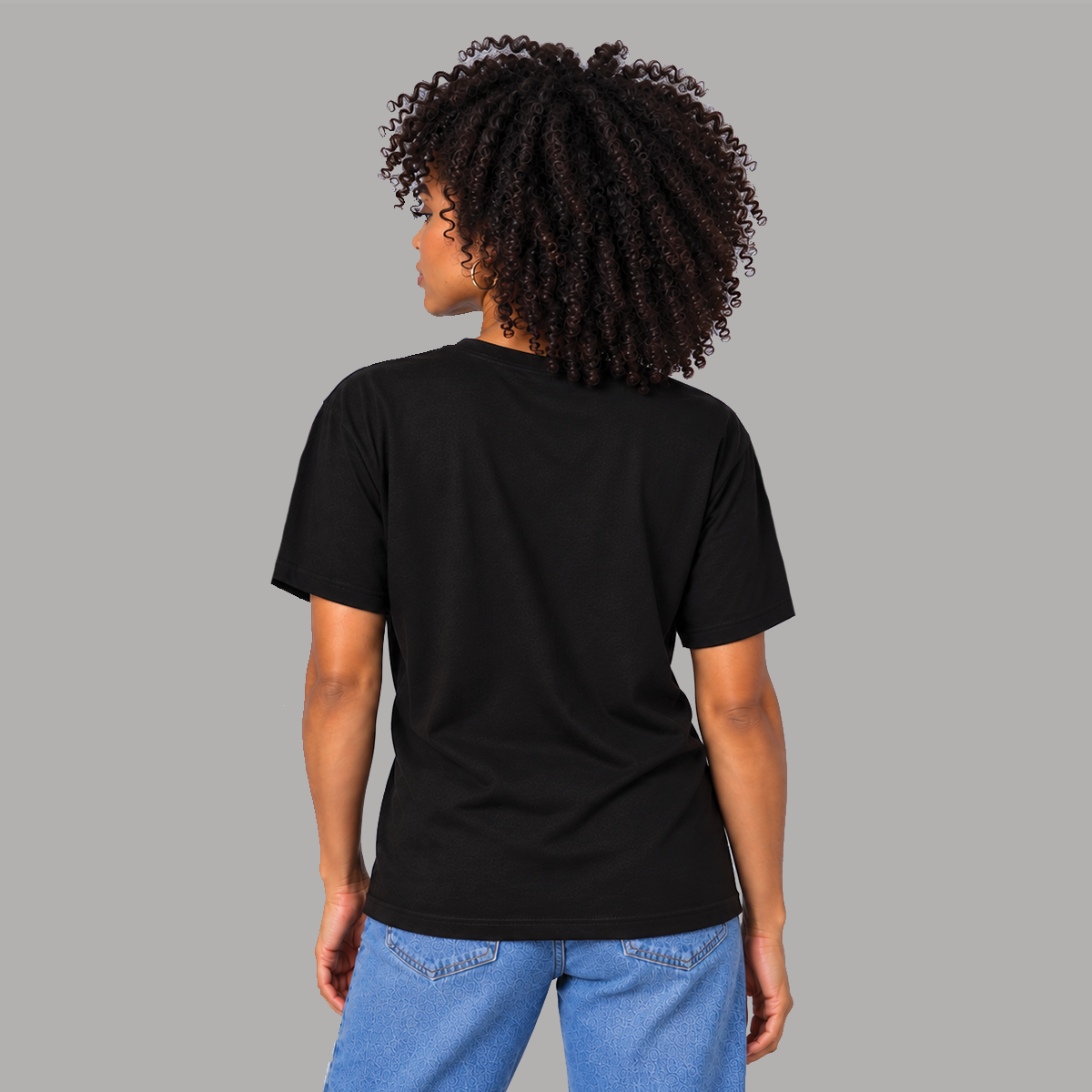 A woman with curly dark hair is shown from the back wearing a plain black short-sleeve t-shirt showing a unisex fit and that there are no graphics on the back.