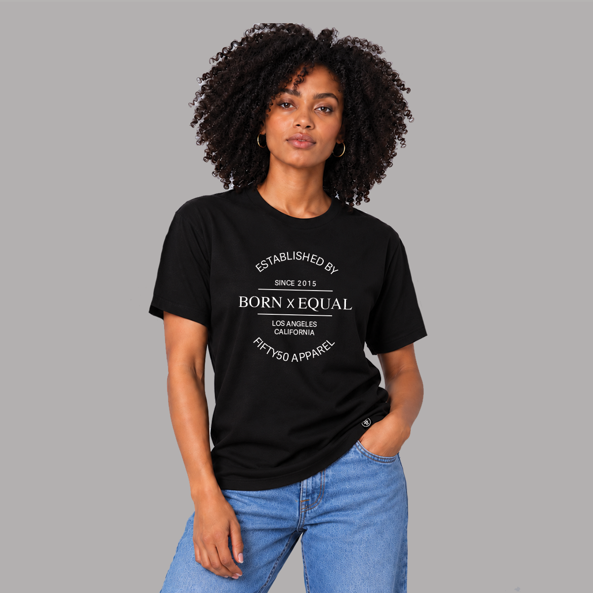 A woman with curly black hair models a black short-sleeve graphic t-shirt with white text that reads 'ESTABLISHED BY BORN x EQUAL LOS ANGELES CALIFORNIA FIFTYSO APPAREL'. She is wearing light wash blue jeans against a solid gray studio background.