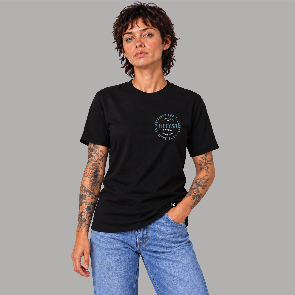 A woman wearing a black crew-neck t-shirt with a circular graphic on the left chest, that says 'DESIGNED IN CALIFORNIA, ESTABLISHED FOR EQUALITY, FIFTY50 APPAREL' paired with blue jeans, standing against a solid gray background.