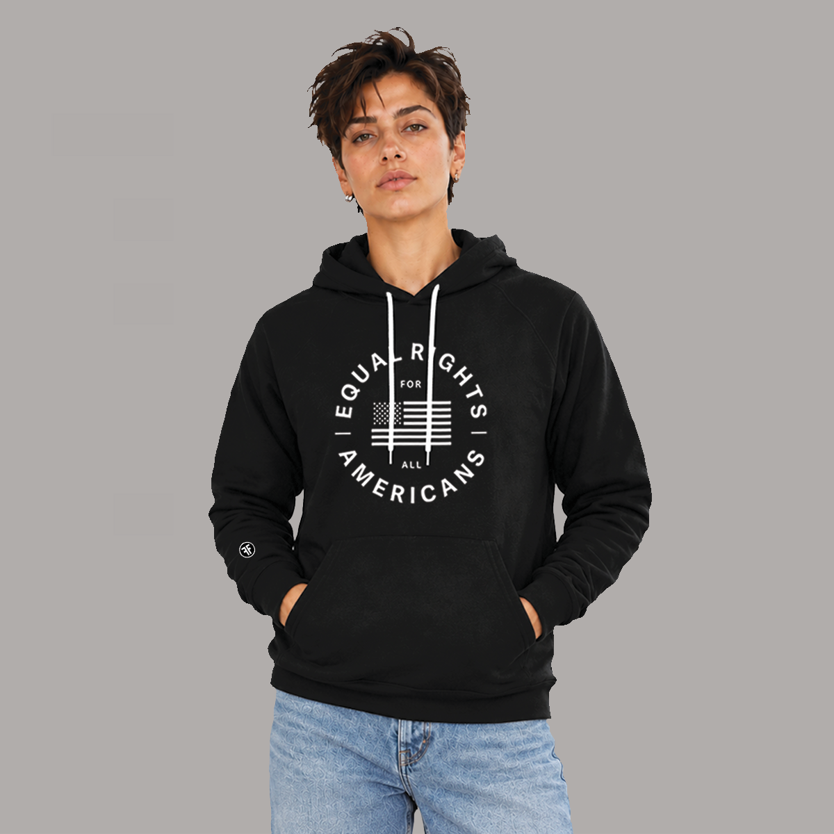 A woman wearing a black pullover hoodie with a white circular graphic on the chest that reads 'EQUAL RIGHTS FOR ALL AMERICANS' around an American flag, paired with light blue jeans, against a solid gray background.