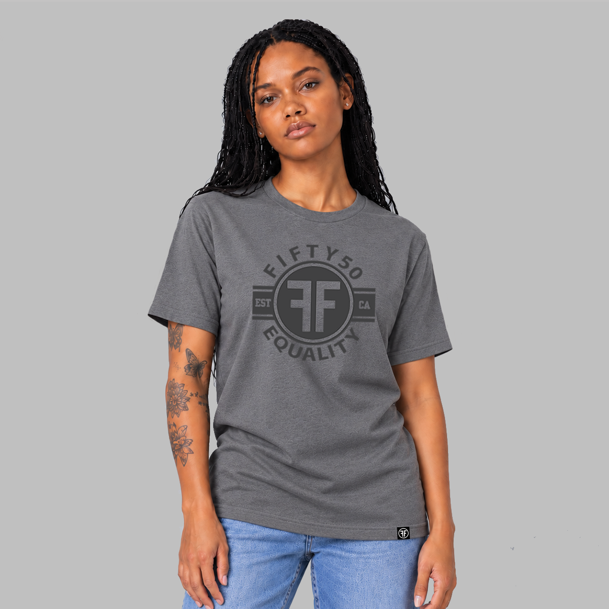 A woman modeling a dark gray crewneck t-shirt with a black graphic on the chest that reads 'FIFTYFO EQUALITY' with a stylized 'FF' logo. She is wearing blue jeans against a solid gray studio background.