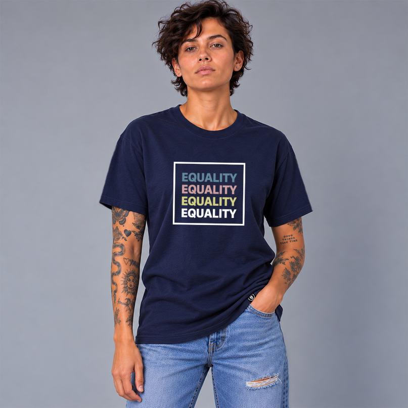 A woman wearing a navy blue graphic t-shirt with the word 'EQUALITY' repeated in a white square box in slate blue, pink, yellow, and white, paired with light-wash distressed jeans, against a solid gray studio background.