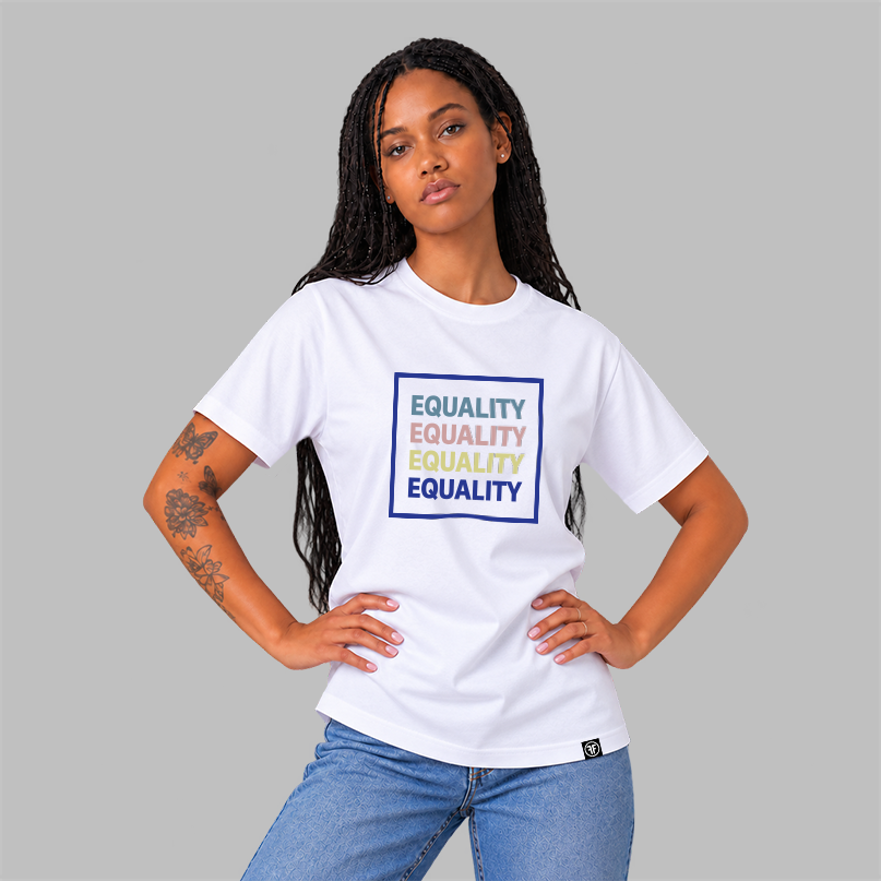 A woman modeling a white short-sleeve graphic t-shirt with the word 'EQUALITY' repeated in slate blue, pink, yellow, and navy blue, inside a navy blue-bordered box, paired with blue jeans against a solid gray studio background.
