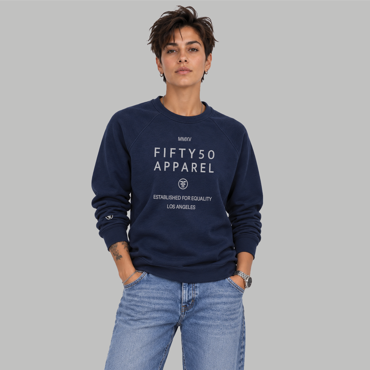 A woman with short hair wearing a navy blue crewneck sweatshirt with a light grey printed text reading 'FIFTY50 APPAREL ESTABLISHED FOR. EQUALITY LOS ANGELES' paired with light wash blue jeans, against a solid gray background.