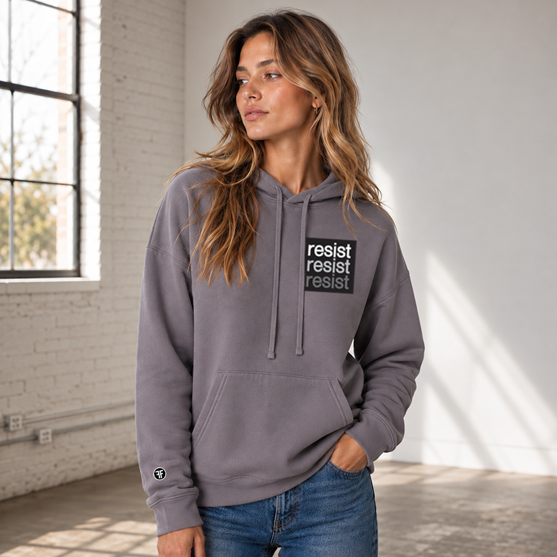 A woman wearing a slate purple pullover hoodie with a black chest logo that reads 'resist resist resist' in white, light grey, and dark grey text, paired with blue jeans, against a solid gray background.