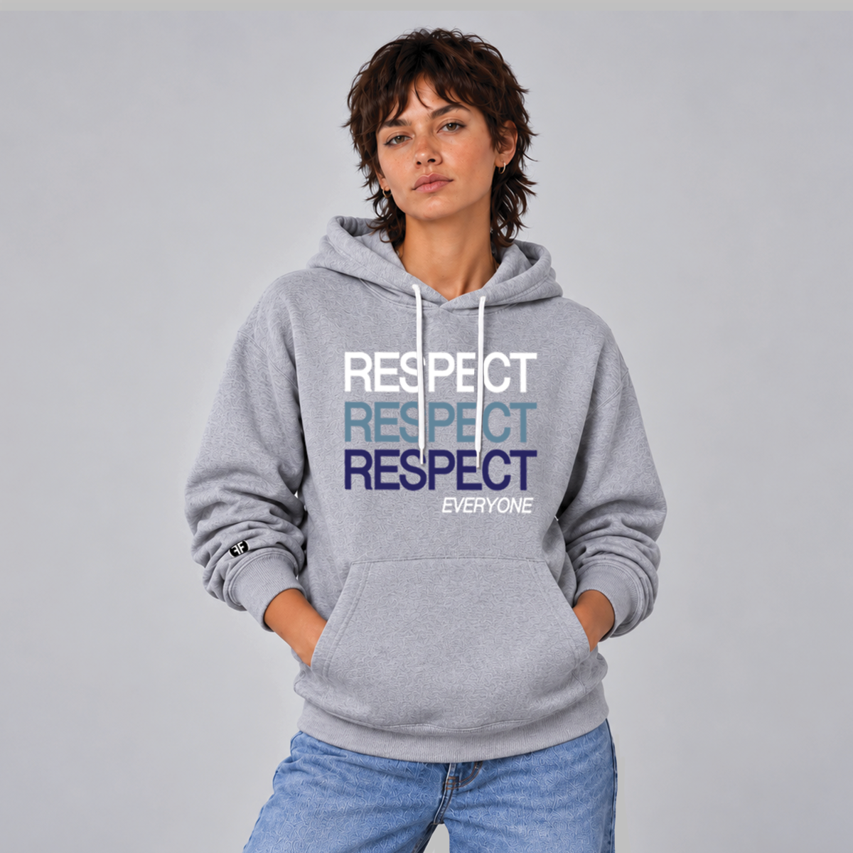 A woman modeling a heather gray pullover hoodie with a graphic text print reading 'RESPECT RESPECT RESPECT EVERYONE' in white, light blue, and dark blue, paired with blue jeans against a solid gray studio background.