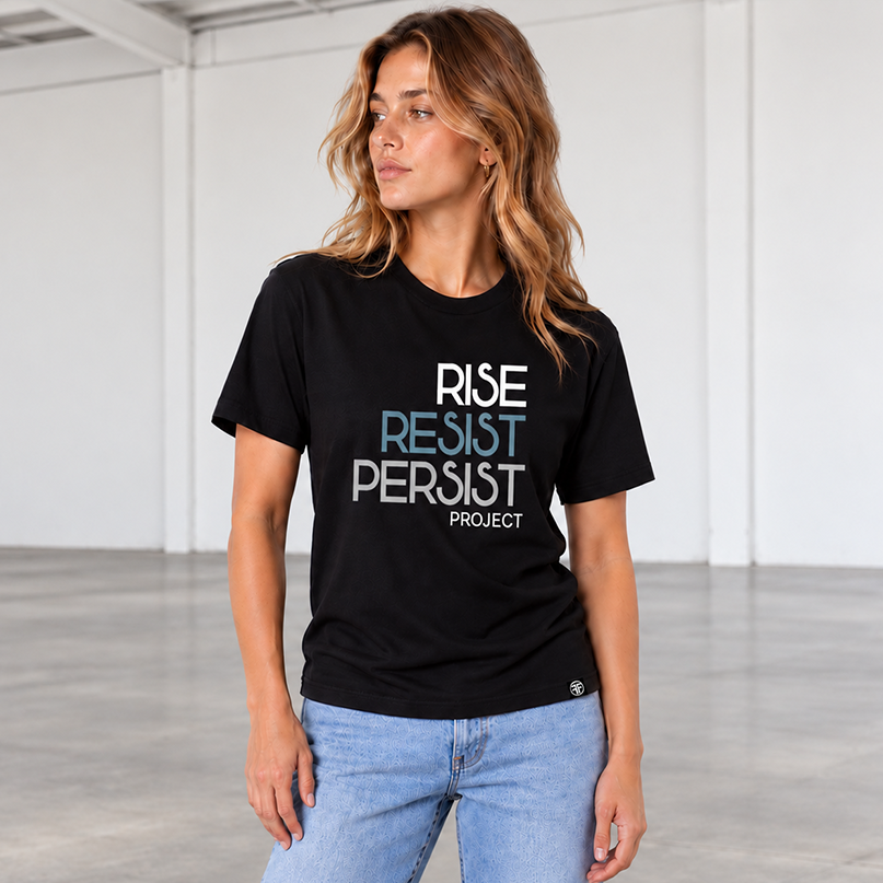 A woman wearing a black graphic t-shirt with the text 'RISE RESIST PERSIST PROJECT' in white, stone blue, and light grey paired with light blue jeans, standing in a bright, modern studio with white walls and a gray concrete floor.
