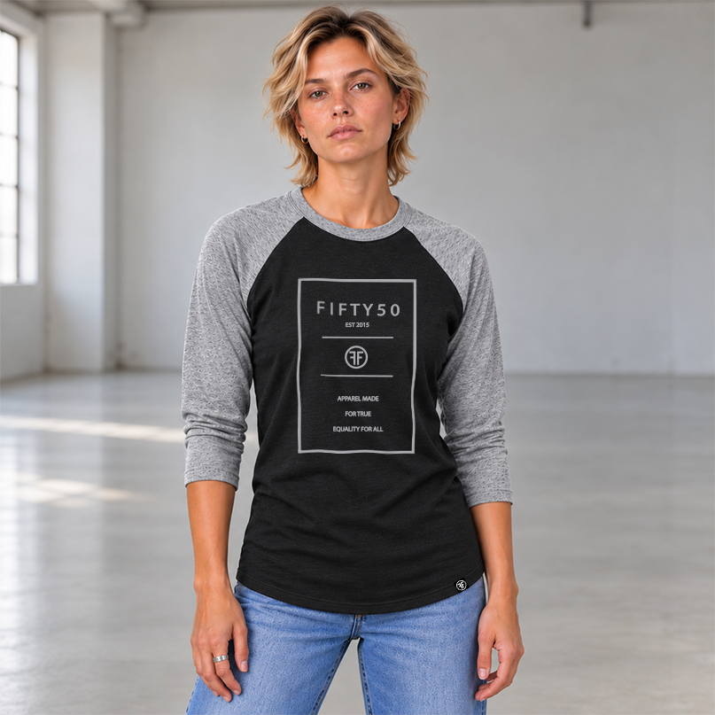 A lady wearing a 3/4 sleeve heather black baseball t-shirt with heather grey arms and collar. The logo is a grey rectangle with the grey text Fifty 50, and Apparel Made For True Equality for All inside of it.