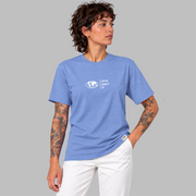 A man wearing a  blue short-sleeve t-shirt with a white graphic center chest featuring a globe and the text 'FIFTYT50 EQUALITY CO. Paired with white pants, set against a solid gray background.